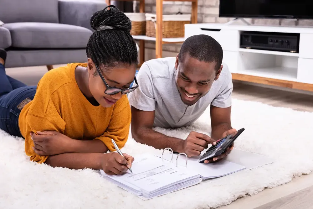 Two people lying on a living room rug reviewing paperwork and using a calculator for household budgeting and financial planning.