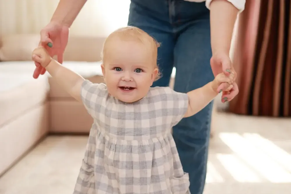 Parent helping a baby take first steps at home, representing early childhood milestones and family planning.