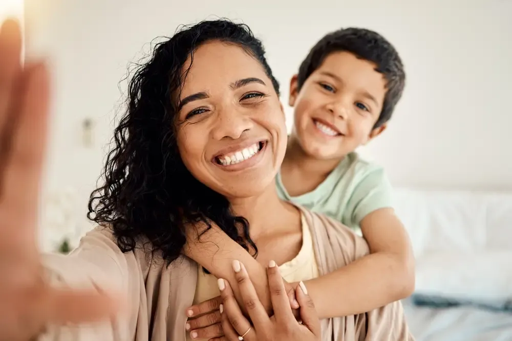 Smiling parent taking a selfie at home while a young child hugs from behind, representing family life and Canada Child Benefit support for parents and children.