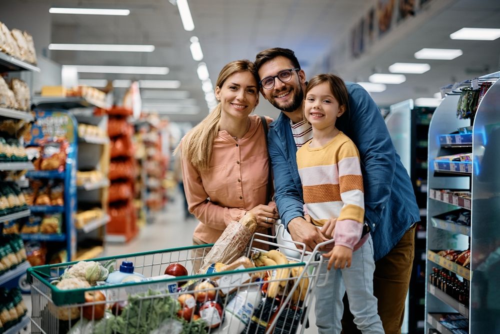 A family grocery shopping together in a supermarket aisle with a full shopping cart filled with fresh produce and household items.