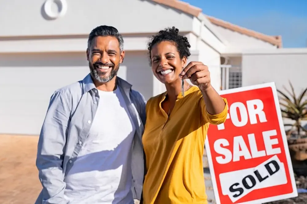 Homeowners standing in front of a sold property sign after selling a house, illustrating a real estate sale that may result in capital gains tax in Canada.
