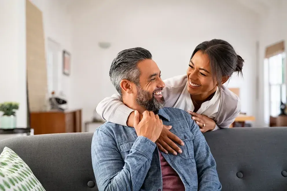 Two adults sitting on a couch in a bright living room, one leaning over and placing arms around the other’s shoulders in an affectionate pose, modern home interior with soft natural lighting.