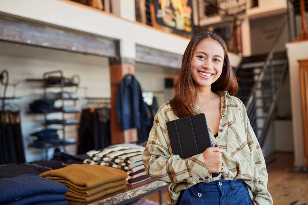Person standing in a clothing store holding a tablet, with folded sweaters on a table and racks of apparel in the background.