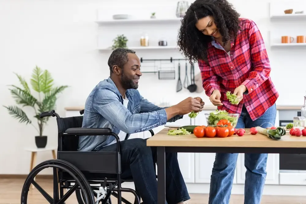 Person using a wheelchair and another person preparing a fresh salad together in a modern kitchen with vegetables on the table.