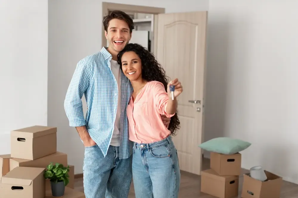 Couple standing in their new home holding a house key with moving boxes stacked around them.