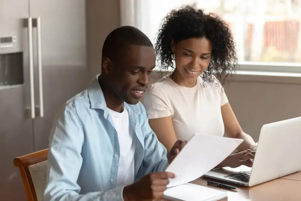 Two people reviewing documents at a table while using a laptop, with papers spread out in front of them in a home setting.