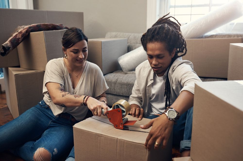 Two individuals sealing a cardboard moving box with packing tape while surrounded by stacks of boxes and moving supplies in a home setting.
