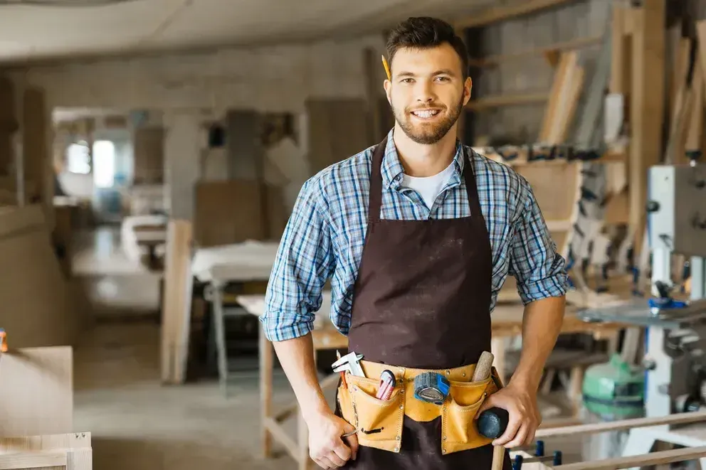 Skilled woodworking professional wearing a tool belt and apron standing in a well-equipped carpentry workshop with woodworking tools and machinery in the background.
