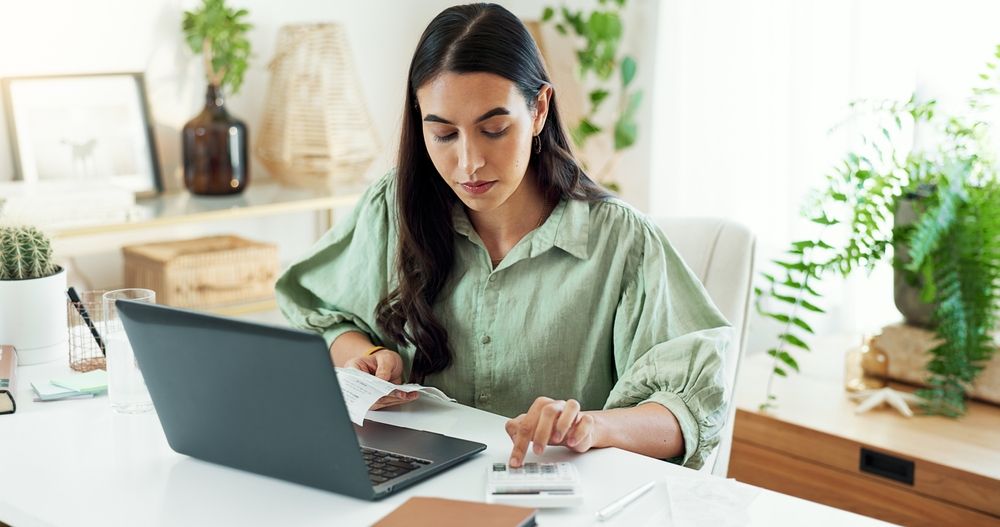 A person working at a desk using a laptop and calculator to review financial documents in a bright home office with plants in the background.