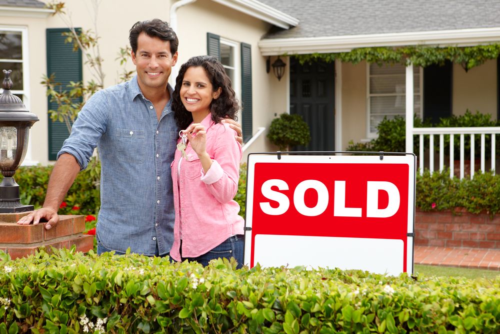 Two people standing in front of a home next to a large ‘SOLD’ sign, holding a key and surrounded by green hedges and a front porch.
